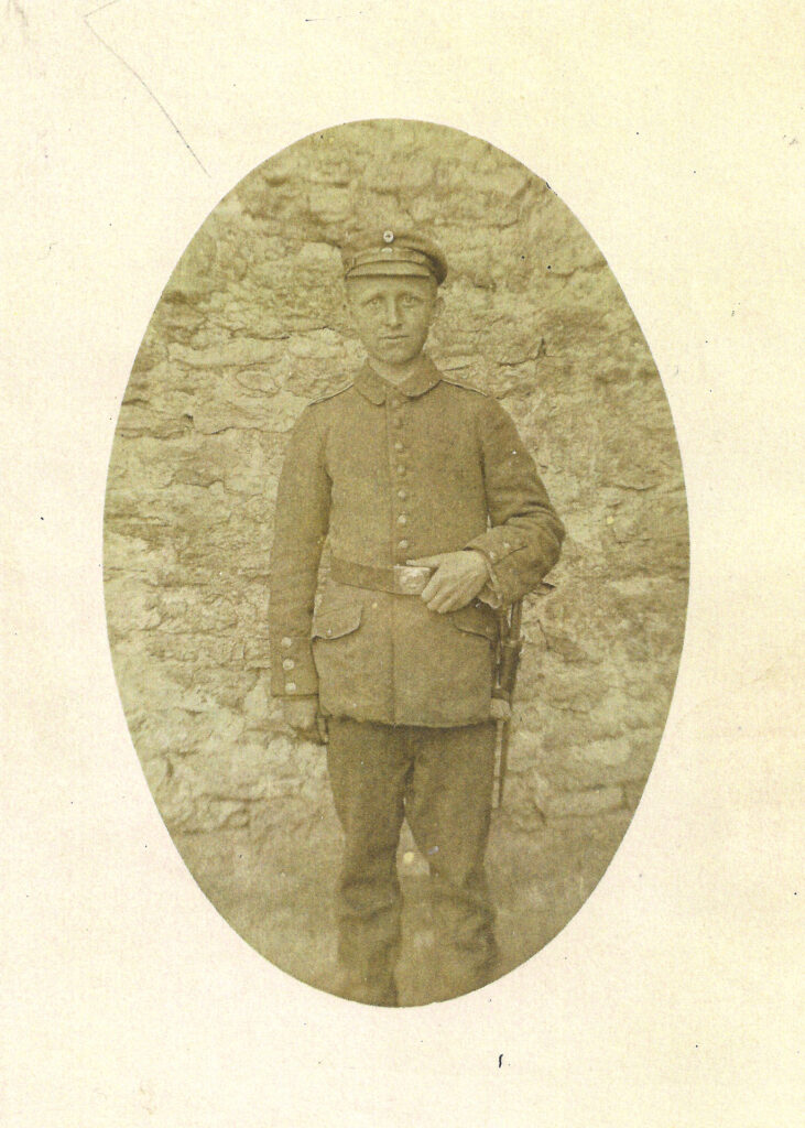 Das vergilbte schwarz-weiße Porträt zeigt Johannes Müller in Uniform. Die linke Hand liegt am Gürtel und er blickt frontal in die Kamera. Er steht vor einer groben teilweise verputzten Steinmauer. Das Foto hat eine ovale Form und ist in ein Passepartout eingefasst.