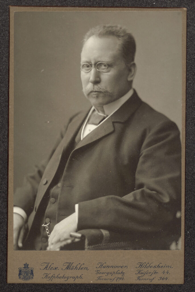 Es ist ein schwarz-weißes Porträt von Carl Wolff. Er trägt einen dunklen Anzug mit Weste, darunter ein helles Hemd. An einem Knopf der Weste ist eine Uhrenkette erkennbar. Er trägt eine kleine, runde Nickelbrille und einen Oberlippenbart. Er hat eine hohe Stirn und "Geheimratsecken".