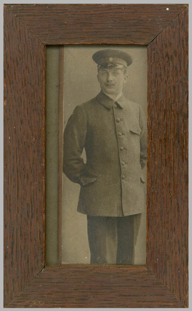Es ist ein schwarz-weißes Foto in einem Holzbilderrahmen. Es ist eine Halbtotale von Wärter Ernst Bünger, sodass er nur bis zu den Knien zu erkennen ist. Er trägt seine Uniform mit Schirmmütze und gestärktem Hemd. Er steht aufrecht, die Hände hinter dem Rücken verschränkt. Er blickt ernst direkt in die Kamera. Er hat einen Schnauzbart.