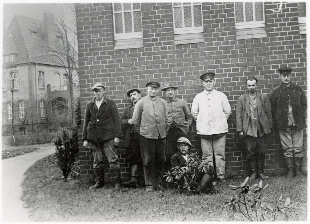 Es ist ein schwarz-weißes Foto. Sechs Erkrankte unterschiedlichen Alters stehen in Arbeitskleidung vor einer Backsteinmauer des Krankenpavillons auf Gut Wienebüttel. Bis auf einer, tragen sie verschiedene Mützen und Hüte, grobe Stiefel. Einer sitzt mit einer Pfeife im Mund im Schneidersitz hinter einem kleinen Rhododendronbusch, der sich vor den Männern befindet. Hinter ihm steht ein Wärter. Er trägt eine helle Uniformjacke und seine Wärtermütze. Einige lächeln in die Kamera.