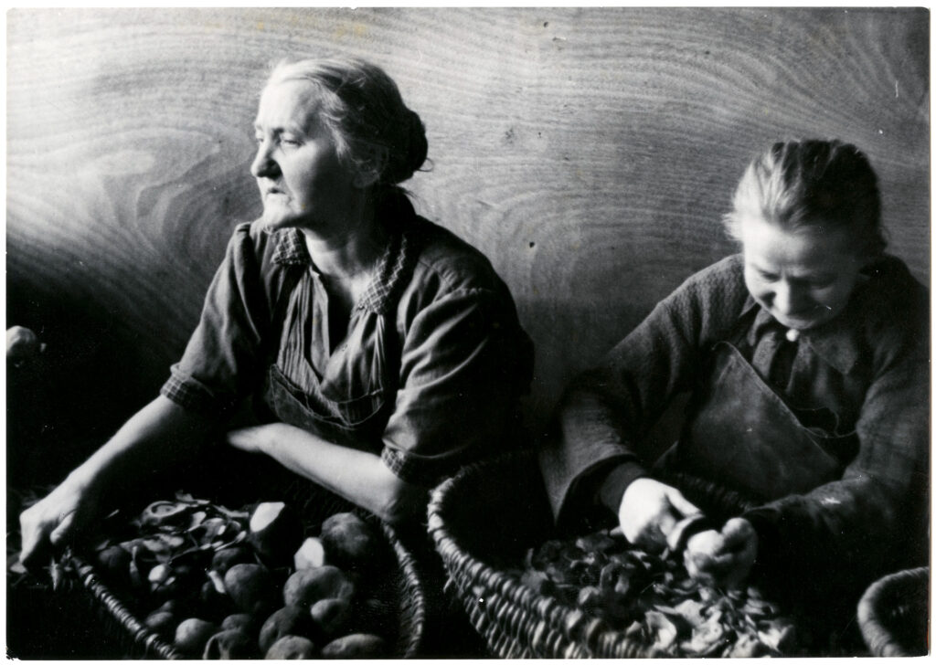 Es ist ein schwarz-weißes Foto von zwei älteren Frauen beim Kartoffelschälen. Sie haben ihre ergrauten Haare zu einem Knoten zurück gebunden. Die Frauen sitzen und haben jeweils einen großen Korb mit Kartoffeln und der abgeschälten Schale auf ihrem Schoß. Sie tragen Arbeitsschürzen. Die Frau rechts im Bild ist auf das Schälen konzentriert, die andere Frau blickt ermüdet zur Seite.