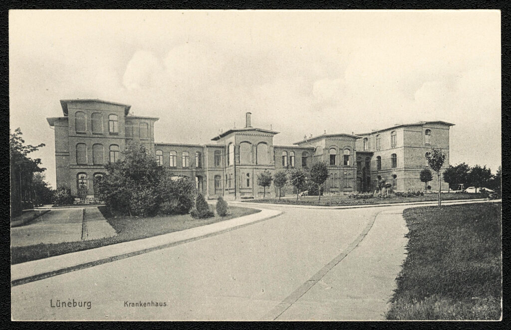 Es ist eine Postkarte mit einem schwarz-weißen Foto. Es ist die Vorderansicht des Städtischen Krankenhauses. Es ist ein breites Backsteingebäude. Die äußeren Trakte sind dreigeschossig, die nach innen liegenden Trakte sind zweigeschossig. Zum Haupteingang des Gebäudes führt eine breite Straße mit Gehsteigen auf beiden Seiten. Vor dem Gebäude sind Bäume frisch angepflanzt.