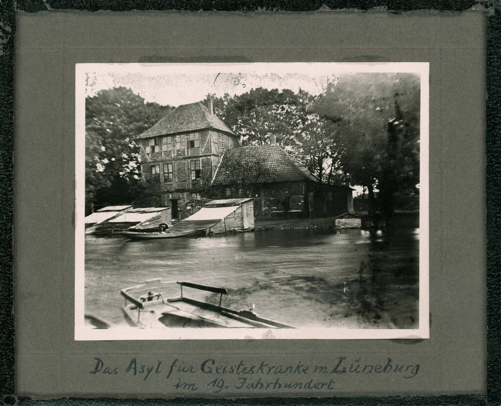 Es ist ein schwarz-weißes Foto. Es hat eine weiße Umrandung. Das Foto zeigt ein zweigeschossiges Fachwerkhaus mit eingeschossigem Seitenanbau. Es steht am Flussufer des Flusses Ilmenau im Wandrahmpark. Im Fluss sind Holzbauten erkennbar und Kähne. Der Fotograf muss auf einem der Kähne gestanden haben, um das Bild vom Wasser aus zu fotografieren.