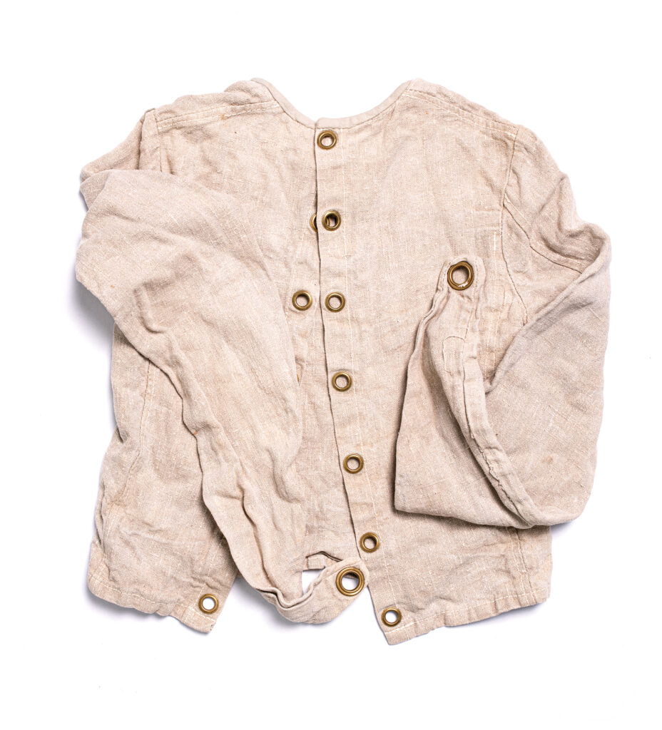 In der Vitrine sind eine Zwangsjacke und ein Fixiergurt. Die Zwangsjacke ist aus einem groben, hellgrauen Leinen genäht. Vor der Brust sowie an den überlangen Ärmeln ist sie mit großen Ösen aus Metall versehen. Die Zwangsjacke liegt gefaltet. Der Fixiergurt ist aus hellbraunem Leder. Er enthält Ösen. Einige Bereiche des Gurtes sind ausgepolstert.