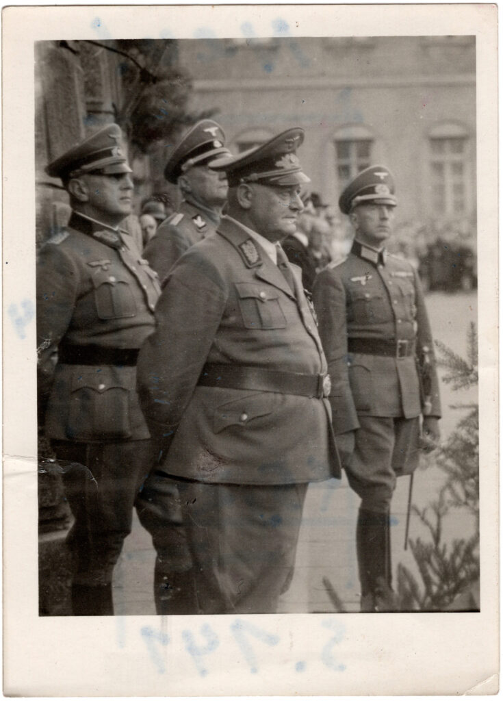 Es ist ein schwarz-weißes Foto von vier Männern in Uniform mit hohen Lederstiefeln und einem breiten Gürtel um die Bäuche. Sie tragen eine Uniformmütze und sie bekleiden verschiedene Orden. Der vorderste, stark gewichtige Mann ist Otto Telschow. Er, und der Mann hinter ihm haben die Hände verschränkt auf dem Rücken. einer trägt Handschuhe. Sie stehen vor der Fassade des Lüneburger Rathauses. Das Bild zeigt sie von der Seite. Am unteren Bildrand sind Tannenzweige.