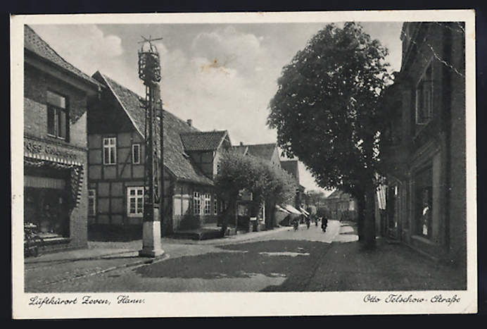 Es ist eine schwarz-weiße Postkarte mit einem weißumrandeten Foto von der Otto-Telschow-Straße in Zeven. Die Straße ist auf der einen Seite mit hohen Bäumen gesäumt. Auf der anderen Seite sind zweigeschossige Fachwerkhäuser. An der Seite steht gedruckt: Luftkurort Zeven (Hann.). Otto-Telschow-Strasse.