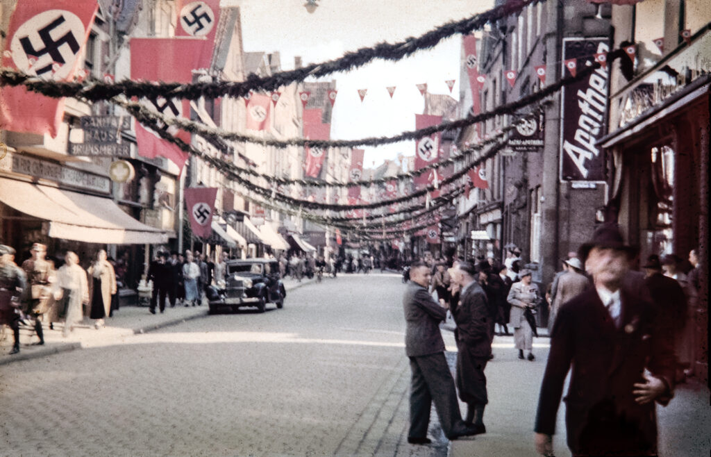 Es ist ein Farbfoto von der Großen Bäckerstraße in Lüneburg. Rechts im Bild ist die Ratsapotheke erkennbar, gegenüberliegend ein Sanitätshaus. In der Mitte verläuft in Kopfsteinpflasterung eine Fahrbahn. Auf jeder Straßenseite gibt es einen schmalen Gehweg, die Straße ist gesäumt von Geschäften. Die Gehwege sind mit Menschen belebt. Auf der Straße parkt ein schwarzes Auto. Zwei Männer stehen halb auf der Straße, einer raucht. Die Straße ist mit Girlanden und Wimpel geschmückt. Überall an den Häuserfassaden hängen große Hakenkreuzfahnen.
