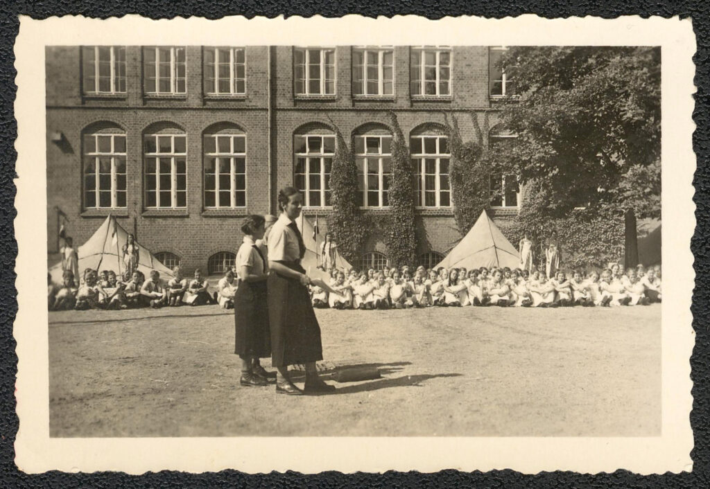 Es ist ein schwarz-weißes Foto. Es zeigt den Schulhof der Wilhelm-Raabe-Schule. Es ist Sommer. Auf dem Hof vor der Gebäudefassade stehen drei große weiße Zelte. Vor jedem Zelt stehen zwei Mädchen. Es sind Fahnen aufgestellt. Auf dem Hof sitzt in einer langen Reihe eine große Gruppe Mädchen. Sie tragen eine Mädchenuniform: ein weißes, kurzärmeliges Hemd, ein weißer Rock und ein dunkles Tuch. Nur wenige Mädchen tragen statt Uniform ein Sommerkleid. Im Vordergrund stehen drei junge Frauen. Sie schreiten die Gruppe ab und tragen ebenfalls eine Uniform. Dazu gehören ein dunkler Rock, ein weißes kurzärmeliges Hemd und ein dunkles Tuch.