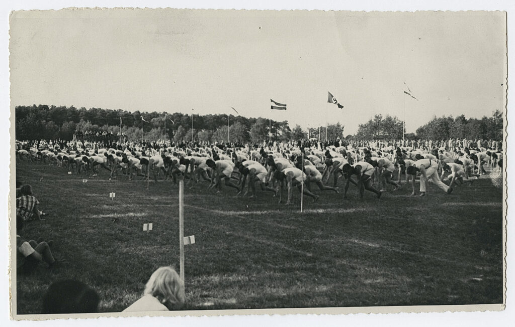 Es ist ein schwarz-weißes Foto. Es zeigt einen Sportplatz. Er ist gefüllt mit hunderten Männern, die in Reih und Glied identische Leibesübungen machen. Sie dehnen ihre Beine. In gebeugter Haltung sind beide Arme auf dem Boden. Zwischen den Händen steht das linke Bein. Das rechte Bein ist nach hinten ausgestreckt. Fast alle Männer tragen weiße kurze Hosen und weiße ärmellose Hemden. Am Bildrand sind Zuschauer erkennbar. Es gibt acht Fahnenmasten, an einem weht die Hakenkreuzfahne.
