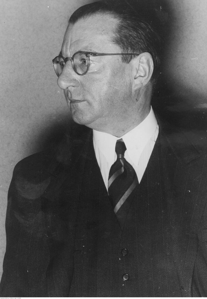 Es ist ein schwarz-weißes Fotoporträt des ältlichen Hellmuth Unger. Er hat eine markante runde Nase, schmale Lippen, die dunklen Haare zurück gekämmt. Er trägt eine Brille und hat Geheimratsecken. Er trägt eine Hornbrille und er hat seinen Kopf zur Seite gedreht und blickt zur Seite. Er trägt ein weißs Hemd, bis oben zugeknöpft, eine gestreifte dunkle Krawatte und ein dunkles Jackett.