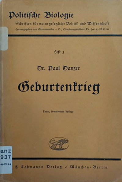 In der Vitrine ist die Originalausgabe des Buches. Das Buch ist geschlossen. Es ist eine Taschenbuchausgabe aus der Schriftenreihe Politische Biologie. Schriften für naturgesetzliche Politik und Wissenschaft. Der Deckel ist aus hellbrauner Pappe. Er ist bedruckt mit dem Titel. Unter dem Titel ist ein Wappen mit einem Löwen gedruckt. Es ist das Wappen des J. F. Lehmann-Verlags.