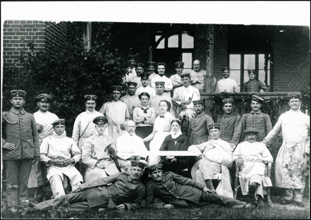 Es ist ein schwarz-weißes Gruppenfoto. Insgesamt 31 Personen stehen auf einer Veranda vor einem Krankenpavillon. Sie blicken in die Kamera. Es sind Pfleger und Pflegerinnen sowie erkrankte Soldaten. Auch eine Nonne und Otto Snell sind unter ihnen. Die Pfleger tragen ihre Uniform mit Mütze und langer, eng geknöpfter Jacke. Die Erkrankten tragen helle Anstaltskleidung und tragen ihr Barrett.