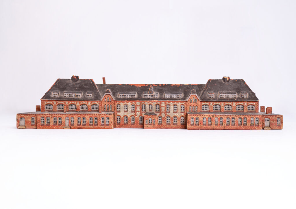 Die Miniatur ist aus Holz. Es ich ein Nachbau des Wirtschaftsgebäudes. Das Gebäude ist langgezogen und besteht aus zwei zweigeschossigen Flügeln. Die Ziegel sind rot, grau und gelb. Über den Fenstern gibt Muster im Mauerwerk. Auffällig ist die reiche Befensterung. Im Erdgeschoss befinden sich zur einen Gebäudeseite im Erdgeschoss 30 Fenster und sechs Türen. Zur gleichen Seite sind über zwei Geschosse Erker mit separatem Eingang. Die Bauart ist von Neunzehnhundert.