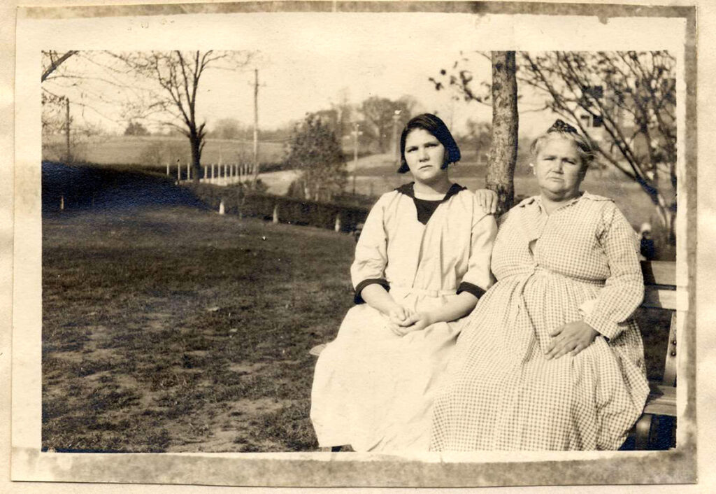 Es ist ein schwarz-weißes Foto. Es hat eine helle Umrandung. Auf dem Foto sitzen Carrie Buck und Ihre Mutter. Carrie sitzt links. Sie hat kinnlange dunkle Haare. Ihre Mutter hat ihre ergrauten Haare hochgesteckt. Beide Frauen tragen helle langärmelige Kleider. Sie sitzen auf einer Parkbank und blicken ernst in die Kamera. Die Mutter hat ihre Hand auf die Schulter ihrer Tochter Carrie gelegt. Im Hintergrund sind Bäume, eine Hecke und eine Straße erkennbar.