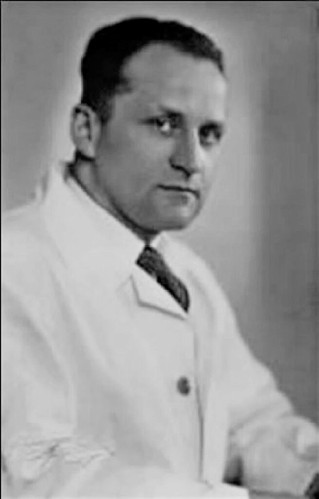 Es ist ein schwarzes-weißes Portraitfoto von Werner Catel. Er trägt eine weißen Arztkittel mit dunkler Krawatte. Seine Haare sind kurzgeschnitten. Er blickt in die Kamera. Sein Blick ist ernst.