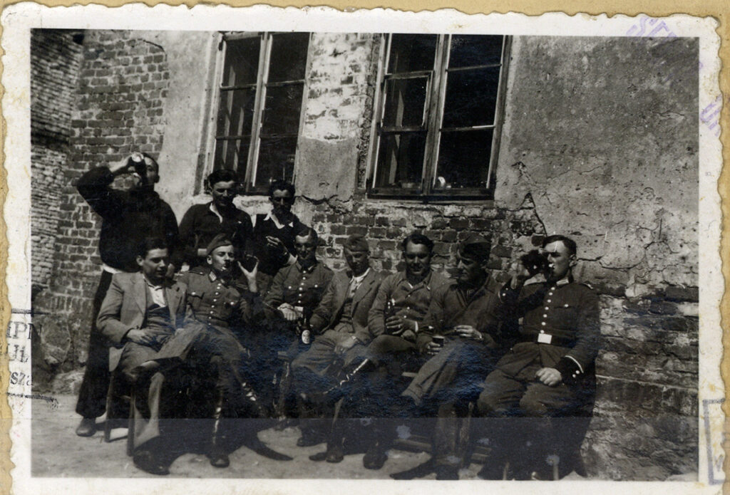 Es ist ein schwarz-weißes Gruppenfoto. Es zeigt sieben Männer in Wehrmachtsuniform, die vor einer Hausfassade, von der der Putz abgeplatzt ist, sitzen und Bier trinken. Hinter ihnen in zweiter Reihe, ebenfalls Bier trinkend, stehen drei Männer. Drei haben die Bierflasche am Mund angesetzt als das Foto entsteht. Die Männer tragen zivile Kleidung, Hemden mit Jacke, Pullover, Pullunder. Die Männer sind jung und mittleren Alters. Es zeigt sie nach der Ermordung von Jüdinnen und Juden mit Gaswagen in Kulmhof.