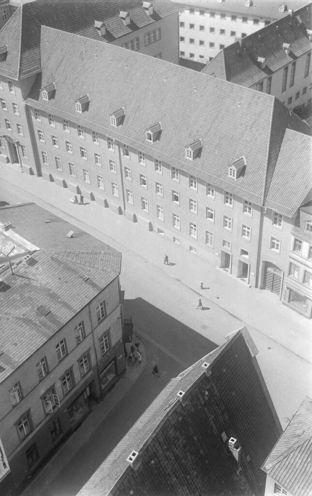 Es ist ein schwarz-weißes Luftbild. Die Aufnahme zeigt den Straßenzug Bardowicker Straße Ecke Lüner Straße in Lüneburg. Es zeigt den Anbau des Gerichtes. Es ist ein langgezogenes dreigeschossiges Gebäude mit einem spitzen Giebeldach. Das Dach hat sechs Gauben mit Sprossenfenstern, eines steht offen. Es stehen noch weitere Fenster offen, die Sonne scheint. Vor dem Gebäude läuft ein Pärchen in Richtung Marktplatz. Der Mann trägt einen Hut und die Frau im kurzärmeligen Kleid schiebt einen weißen Kinderwagen vor sich. Ihnen folgt in etwas größerem Abstand ein weiterer Mann in dunklem Anzug. An der Hausecke Lüner Straße vor dem Kolonialwarengeschäft steht eine Frau mit einem Fahrrad umringt von Kinder. Eine Frau im dunklen Kleid passiert die Straße.