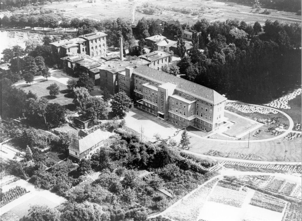Es ist eine schwarz-weiße Luftbildfotoaufnahme. Sie zeigt das Lüneburger Krankenhaus mit dem 1900 eröffneten Hauptgebäude, dem rechten Anbau und den Nebengebäuden. In einem Wäldchen hinter dem Hauptgebäude und dem Heizhaus, erkennbar durch einen großen in den Himmel ragenden Schornstein, erstreckt sich ein langgezogenes Gebäude. Es ist die Baracke. Dahinter verläuft der Oedemer Weg als eine mit Bäumen gesäumte Allee.