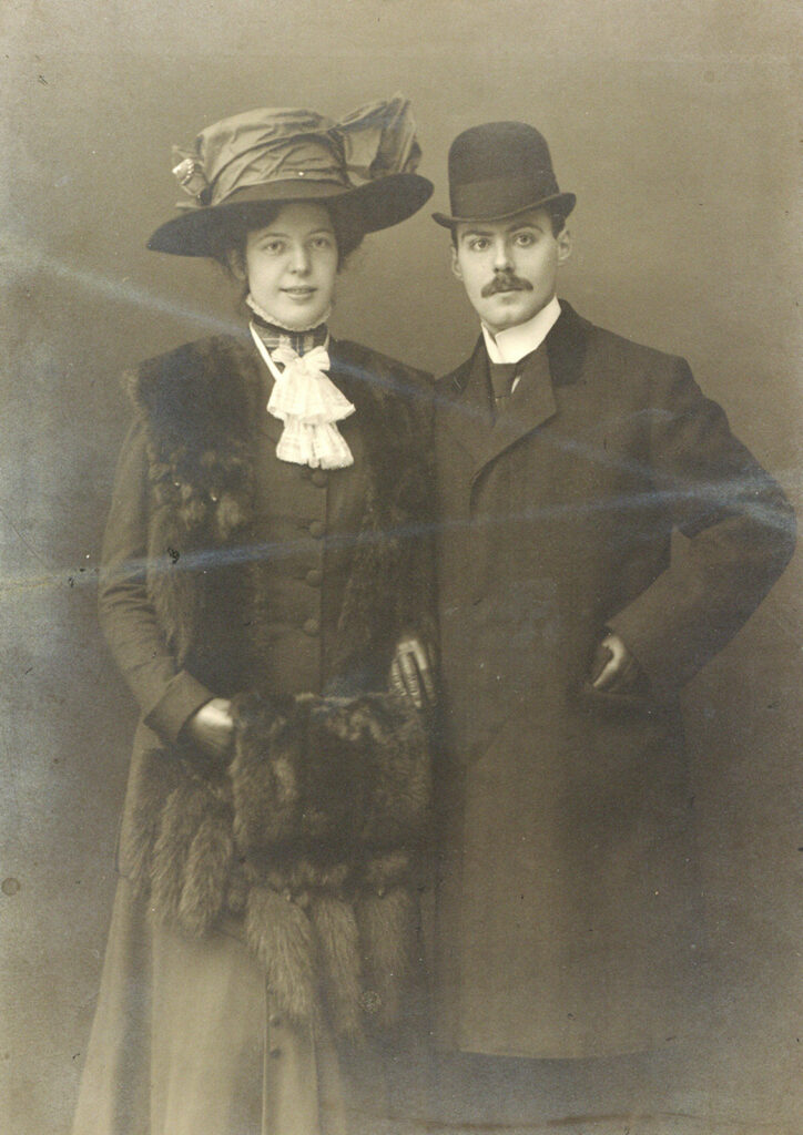 Es ist ein schwarz-weißes Foto von Max Bräuner und seiner Verlobten Helene Feddersen. Helene Feddersen trägt ein über ihrem Dunklen Kleid mit Knopfleiste und Stehkragen, weißem Tuch einen geöffneten Mantel mit einem Pelzbesatz am Revers. Sie trägt Lederhandschuhe und einen Muff aus Pelz. Sie hat eine hochgesteckte Frisur und einen Hut mit großer Schleife und Krempe. Sie lächelt. Rechts neben ihr steht ihr Verlobter Max Bräuner in dunklem, zugeknöpftem Mantel mit abgesetzten Kragen und Melone. Er trägt einen Schnurbart und blickt ernst in die Kamera. Er hat seine linke Hand in der Manteltasche.