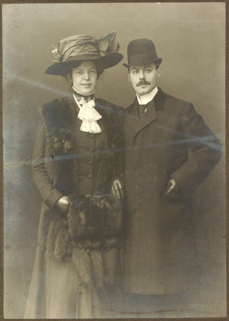 Es ist ein schwarz-weißes Foto von Max Bräuner und seiner Verlobten Helene Feddersen. Helene Feddersen trägt ein über ihrem Dunklen Kleid mit Knopfleiste und Stehkragen, weißem Tuch einen geöffneten Mantel mit einem Pelzbesatz am Revers. Sie trägt Lederhandschuhe und einen Muff aus Pelz. Sie hat eine hochgesteckte Frisur und einen Hut mit großer Schleife und Krempe. Sie lächelt. Rechts neben ihr steht ihr Verlobter Max Bräuner in dunklem, zugeknöpftem Mantel mit abgesetzten Kragen und Melone. Er trägt einen Schnurbart und blickt ernst in die Kamera. Er hat seine linke Hand in der Manteltasche.