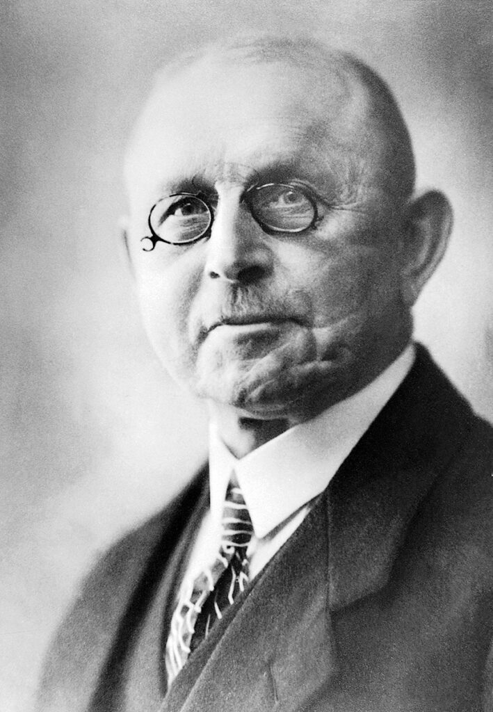 It is a black-and-white portrait of Heinrich Behr. He has a trimmed moustache and wears rimless nickel-rimmed glasses. He is wearing a dark suit, shirt and striped tie. He has hardly any hair left. His face is furrowed by two scars.