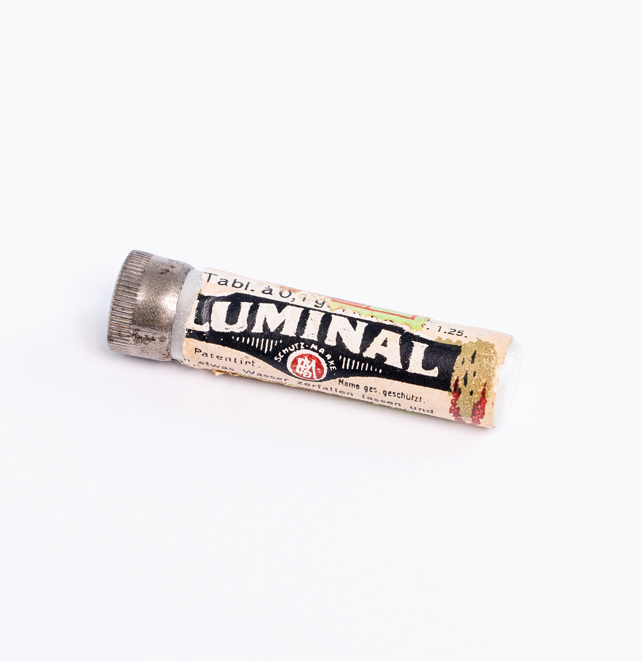 Die Dose ist ein mit einem Schraubdeckel verschlossenes Röhrchen. Es trägt ein Etikett, auf dem mit weißer Schrift auf schwarzem Untergrund in großen Buchstaben steht. Auf dem Etikett steht: Tabl. a 0,1g.