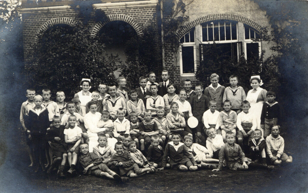 Es ist ein schwarz-weißes Gruppenfoto von 40 Jungen im Alter von sechs bis 16 Jahren, drei Pflegerinnen und zwei jungen Männern. Viele Jungen tragen Matrosenhemden und einen Pullover, kurze Hosen, Strümpfe und Stiefel. Die älteren Jungen tragen Hemden und Jacken. Eine Pflegerin sitzt inmitten der Gruppe und hat einen Jungen auf dem Schoss. Zwei Pflegerinnen tragen ihre Uniform mit Schwesternhaube. Die Gruppe steht in vier Reihen im Garten eines der Gebäude der Heil- und Pflegeanstalt Lüneburg. Die meisten Jungen lachen in die Kamera.