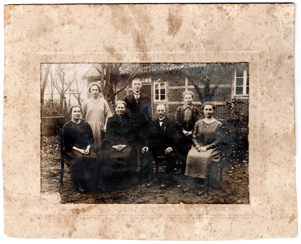 Es ist ein schwarz-weißes Gruppenfoto in einem Passepartout. Vier Familienmitglieder sitzen auf Stühlen in der ersten Reihe. Drei Familienmitglieder stehen in zweiter Reihe dahinter. Sie haben sich vor der Fassade des Fachwerkhauses des Hofes aufgebaut. Alle tragen festliche Kleidung und blicken ernst in die Kamera.