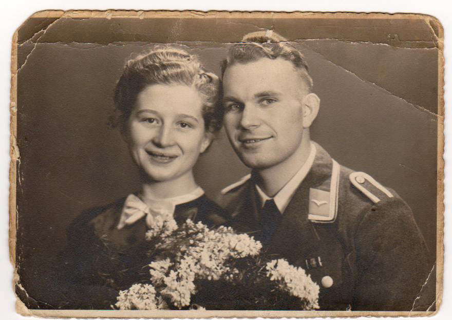 Es ist ein schwarz-weißes Foto. Erich und Thea blicken lächelnd in die Kamera und neigen die Köpfe zueinander. Erich trägt Uniform und die Haare zurückgekämmt. Thea trägt ein dunkles Oberteil mit heller Schleife am Hals. Ihre Haare sind gewellt und nach hinten gesteckt. Sie hält einen üppigen Blumenstrauß vor sich.