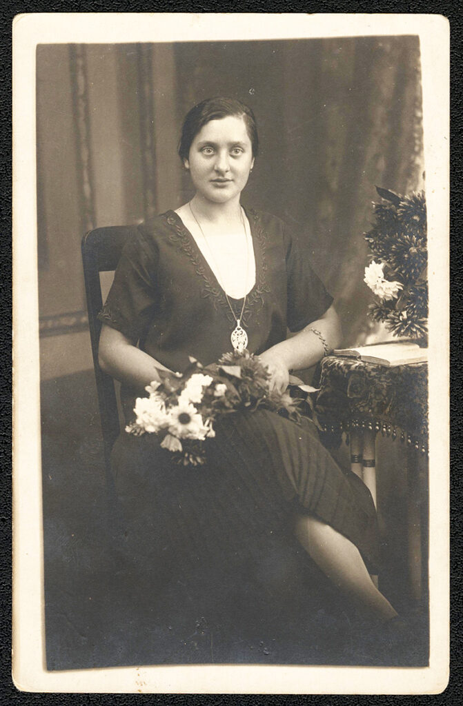 Es ist ein schwarz-weißes Bild. Auguste Schmid sitzt mit verschränkten Beinen auf einem Stuhl und hält einen Blumenstrauß. Sie trägt ein dunkles Kleid mit großem Ausschnitt. Sie trägt eine lange Halskette mit großem, ovalem Anhänger. Sie blickt mit leichtem Lächeln in die Kamera.