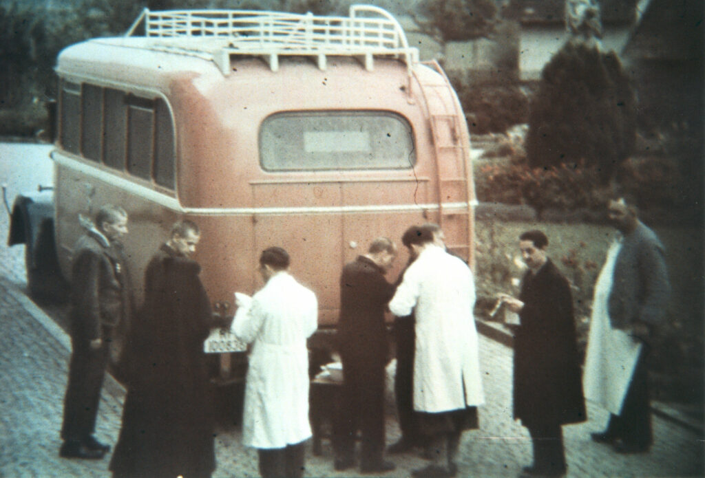 Es ist ein farbiges Foto. Darauf ist ein roter Postbus von hinten zu sehen. Vor dem Bus stehen zwei Ärzte in weißen Kitteln. Außerdem stehen mehrere Männer in dunkler Kleidung bei den Ärzten. Es sind Erkrankte. Die Ärzte kontrollieren ihre Papiere zu den Krankentransporten und prüfen, ob die Erkrankten auf der Liste stehen.