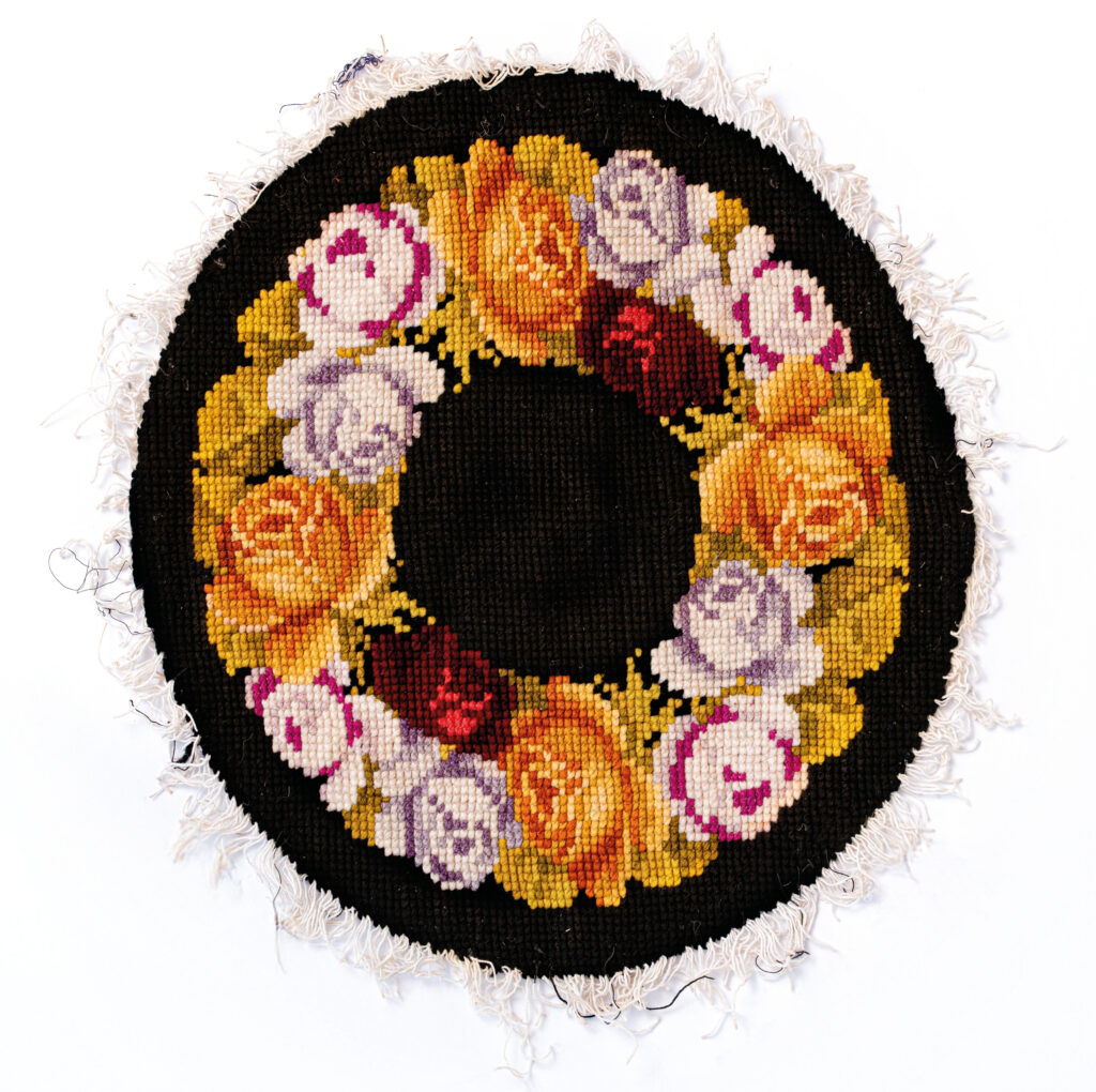 Die Stickerei ist rund. Am Rand sind weiße Fransen. Auf schwarzem Hintergrund ist der Blumenkranz in Kreuzstichstickweise gestickt. Die Blumen sind orange, rosa und rot mit etwas grün.