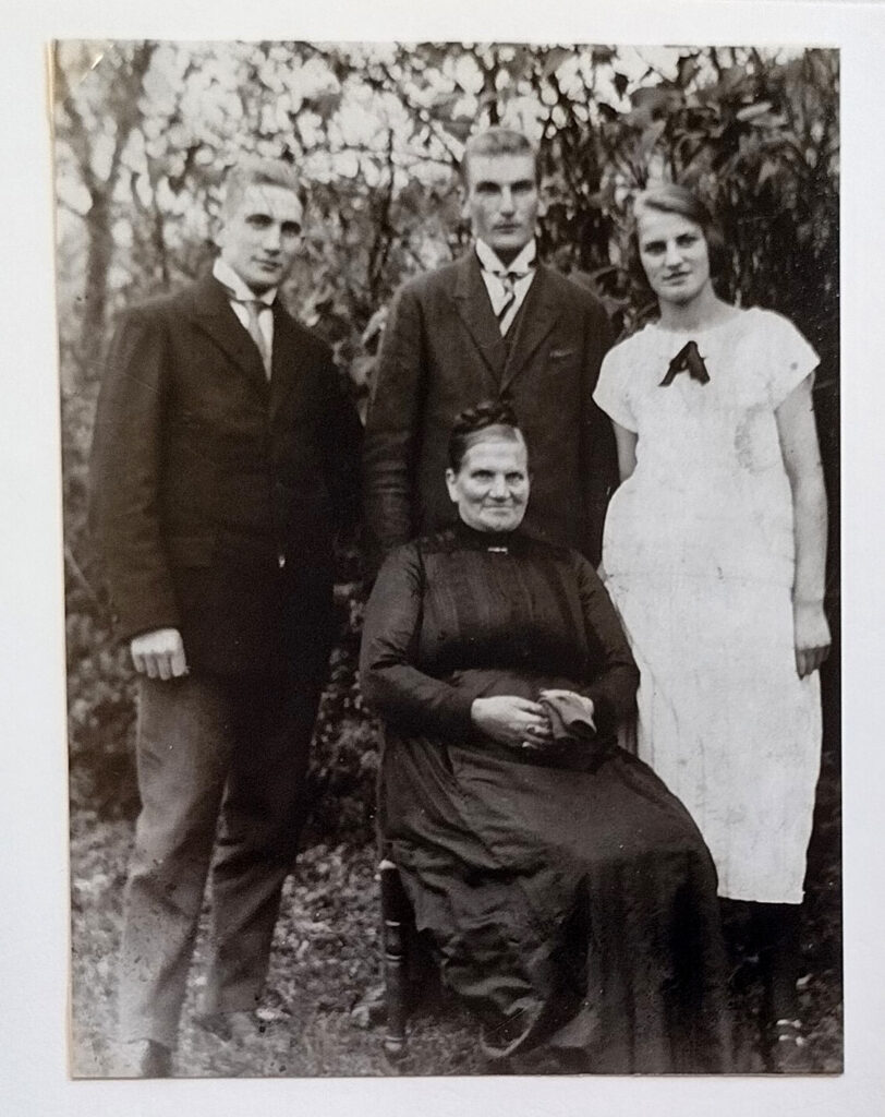 Auf dem schwarz-weißen Familienfoto sitzt die Mutter auf einem Stuhl. Sie trägt ein langes, hochgeschlossenes dunkles Kleid mit streng zurückfrisiertem Haar. Die Hände halten dunklen Stoff im Schoß. Hinter ihr stehen die Geschwister. Die Männer tragen dunkle Anzüge mit Krawatte und hellem Hemd. Alma trägt ein langes weißes Kleid mit kurzen Ärmeln und einer dunklen Schleife am Hals. Alle vier blicken in die Kamera. Im Hintergrund sind Büsche zu erkennen.