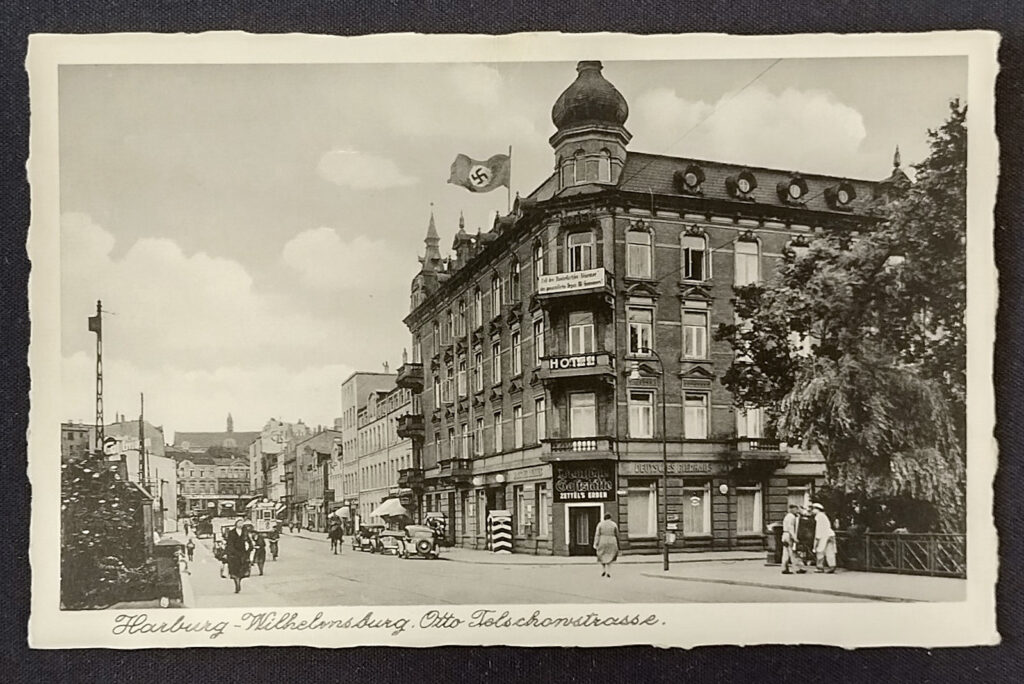 Es ist eine schwarz-weiß-Aufnahme. Das Hotel ist ein vierstöckiges Gebäude an der Ecke einer großen Straße. Es hat einen markanten Zwiebelturm an der Ecke des Gebäudes. Oben auf dem Dach weht eine große Fahne mit Hakenkreuz.