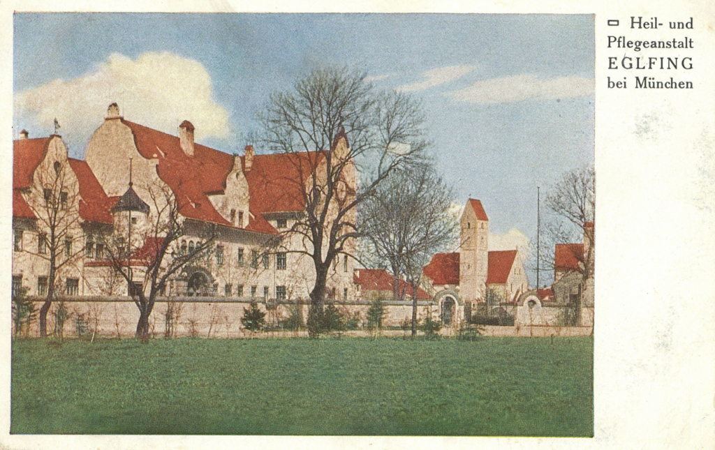 It is a coloured drawing. Surrounded by a light-coloured plastered wall, several light-coloured buildings with red roofs can be seen. On the left in the foreground is a large two-storey house with dormer windows in the roof. Further back on the right is a house with a high tower like a church.