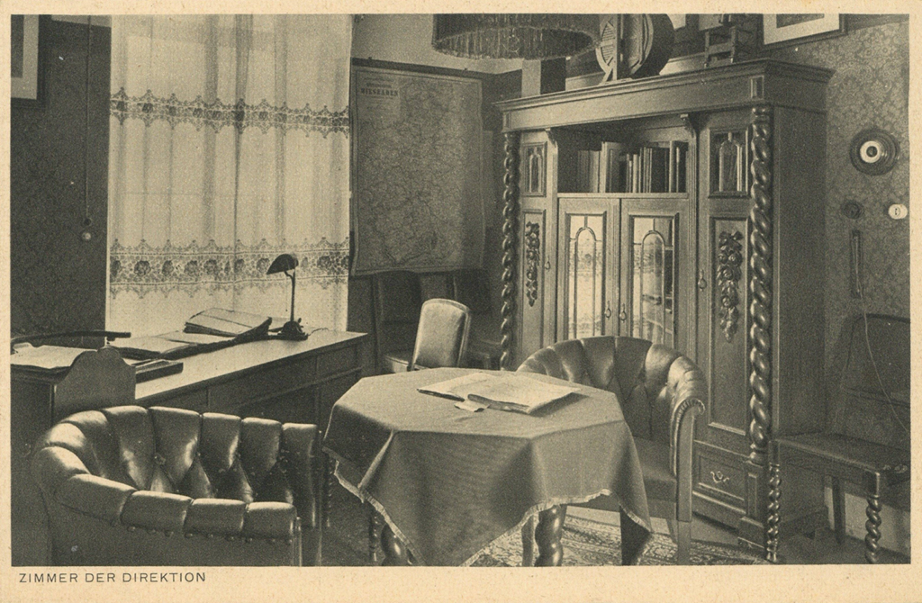 It is a black and white photo. There is a desk in front of a window with a curtain. In front of it is a settee with upholstered chairs. Behind it is a half-height sideboard with leaded glass panes and ornately carved doors.