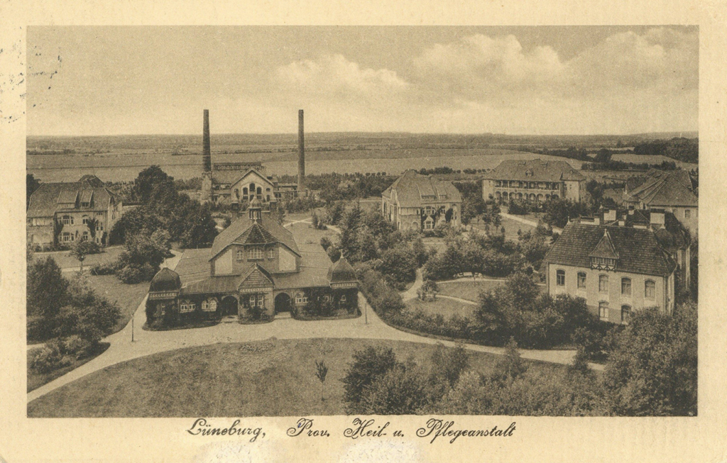 It is a black and white photo. It shows a park-like garden with several villa-like buildings. Fields can be seen at the end of the grounds. Two tall, narrow chimneys can be seen at the far end of the grounds.
