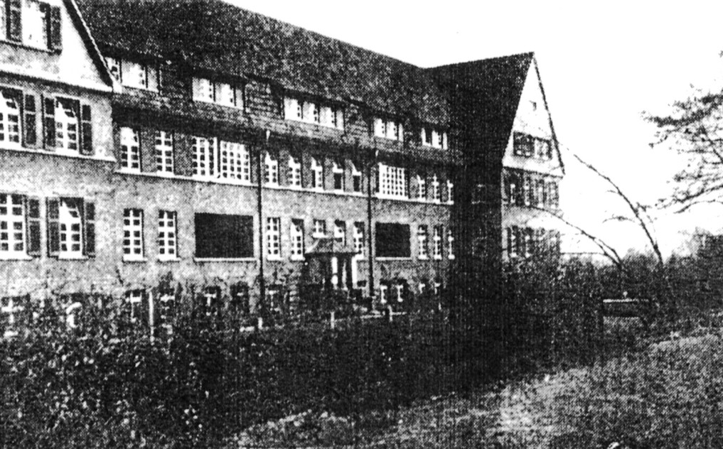 Das Gebäude hat drei Stockwerke und lange Gauben im Dach. Jede Etage hat viele Sprossenfenster. Im ersten Stock sind zwei zurückgesetzte Balkone zu sehen.