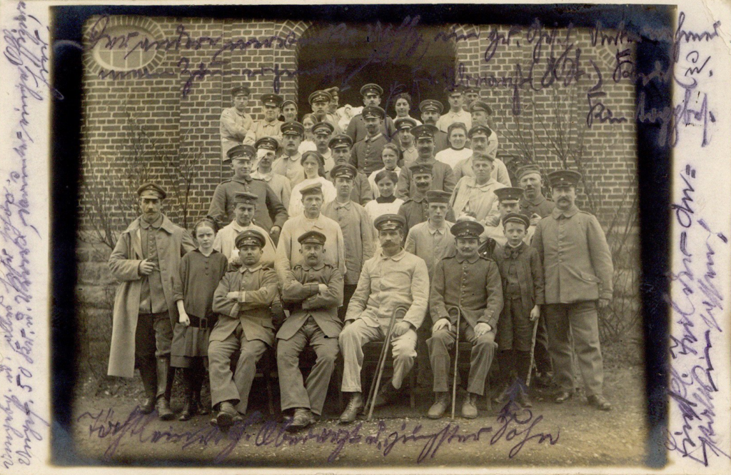 Es ist ein schwarz-weißes Foto. An die Ränder des Bildes sind handschriftliche Notizen eingetragen. Zu sehen ist eine große Gruppe Menschen ordentlich zum Foto aufgestellt. Die Männer und Jungen tragen unterschiedliche Anzüge mit Uniformmützen. Die Frauen tragen Flechtfrisuren und Kittel über den Kleidern.
