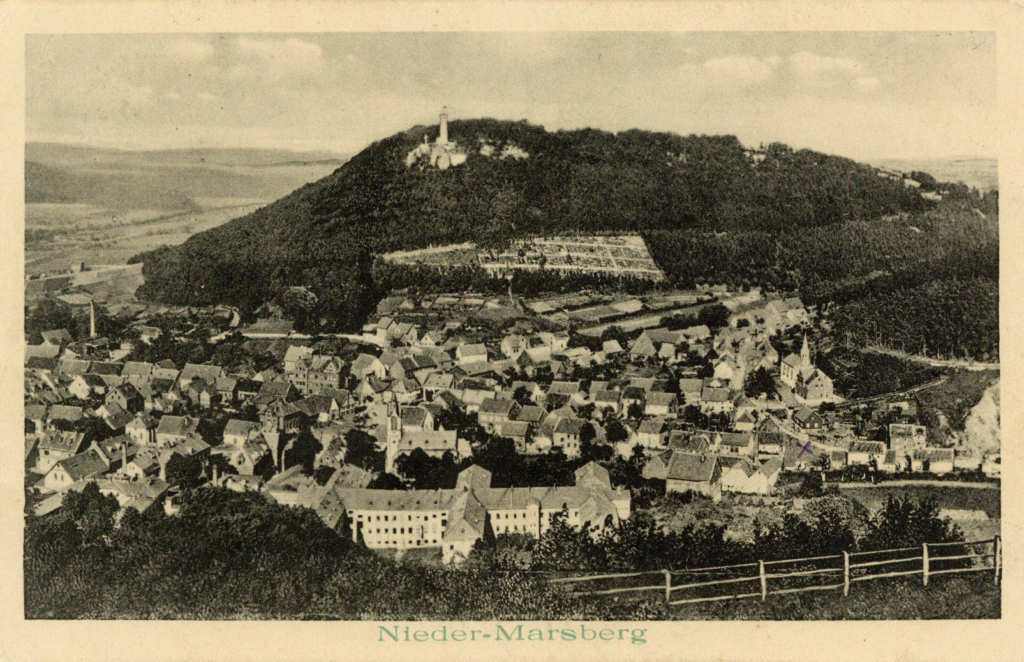 It is a black and white photo. It is a panoramic view from a hill to a valley below. There are settlements in the valley. Behind it is a hill with a high tower in a dense forest.