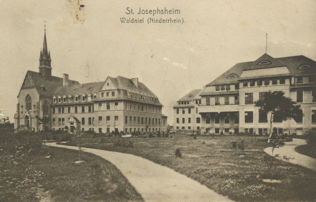 Es ist ein schwarz-weißes Foto. Es sind zwei Gebäude zu sehen. Links steht ein dreistöckiges Haus mit ausgebautem Dach und hohem Giebel. Links am Gebäude ist ein kirchenartig aussehendes Gebäude mit hohem Turm und kunstvollen Fenstern angeschlossen. Rechts Ist ein Gebäude mit vier Stockwerken und daran anschließenden kleineren Gebäuden angebaut. Die Fenster sind hohe Sprossenfenster. Davor liegt eine angelegte Gartenfläche mit anscheinend frisch eingepflanzten Bäumen.