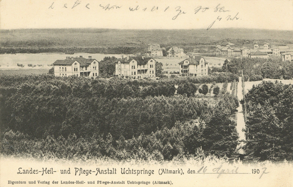 It is a black and white photo. Surrounded by tall woods are several similar buildings. They are three storeys high with dark roofs. The houses have wooden balconies on the sides.