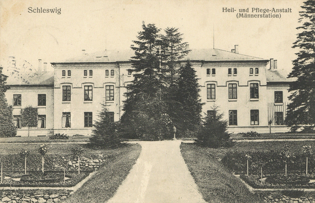 Es ist ein schwarz-weißes Foto. Hinter einer gepflegten Gartenanlage liegt ein helles dreistöckiges Haus. Es hat hohe Sprossenfenster und zwei niedrigere Flügel auf jeder Seite.