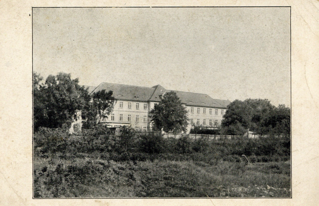 It is a black and white photo. A three-storey, elongated building can be seen. There are many high mullioned windows in the light-coloured façade. There are some trees in front of the building. A light-coloured fence appears to surround the building and the trees. On the other side of the fence and thus in the foreground of the picture are bushes and a kind of natural meadow.