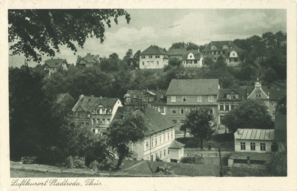 Es ist ein schwarz-weißes Foto. Im Tal und auf einem Hügel sind mehrere Häuser zumeist zweistöckig zu sehen. Zwischen den Häusern sind Gärten zu sehen. Entlang einer Straße in der Mitte des Bildes stehen die Häuser direkt aneinander.