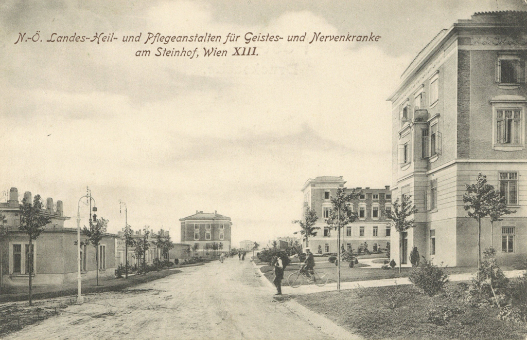 Es ist ein schwarz-weißes Foto. Zu sehen ist ein Blick eine Straße hinunter. Links stehen flache Gebäude mit hohen Sprossenfenstern. Rechts sind zwei vierstöckige Gebäude mit hohen Sprossenfenstern zu sehen. Am Ende der Straße steht ein weiteres solches Gebäude. Auf der Straße sind Personen zu Fuß und auf Fahrrädern zu sehen.