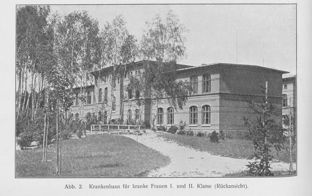 Es ist ein schwarz-weißes Bild. Hinter jungen hohen Birken in einem angelegten Garten ist ein flaches zweistöckiges Gebäude zu sehen. Es hat viele halbrunde Sprossenfenster und ein Flachdach.