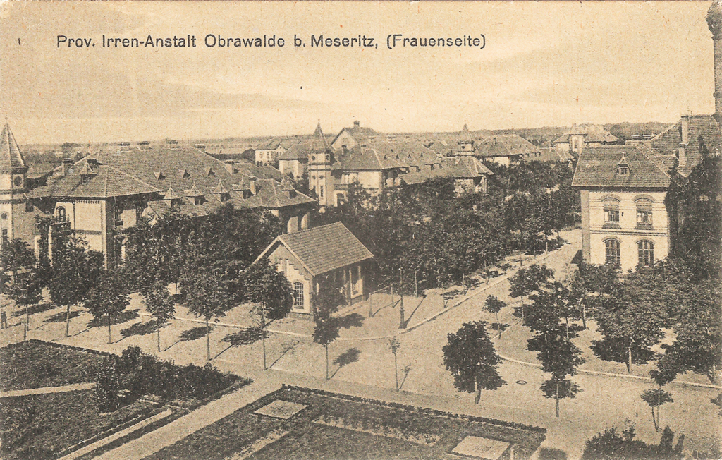 It is a black and white photo. It was taken from a slightly elevated perspective. You can see a fork in the road, with landscaped gardens in the foreground. Behind it is a settlement of many, mostly two-storey houses. These have curved mullioned windows and decorated brick walls. A tall tower is half recognisable on the right-hand edge of the picture, which is a black and white photo. It was taken from a slightly elevated perspective. A fork in the road can be seen, with landscaped gardens in the foreground. Behind it is a settlement of many, mostly two-storey houses. These have curved mullioned windows and decorated brick walls. A tall tower is half recognisable on the right-hand edge of the picture.
