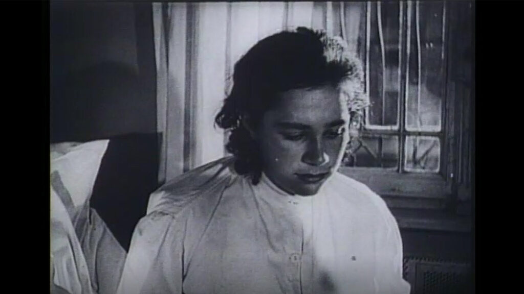 Es ist ein schwarz-weißes Standbild aus dem Film »Dasein ohne Leben«. Die Aufnahme ist unscharf. Sie zeigt eine junge Frau in einem bis zum Hals zugeknöpften Nachthemd, aufrecht sitzende in einem Bett. Das Kopfkissen ist erkennbar. Die leicht zerzausten, braunen Haare sind nach hinten gebunden. Sie hat einen gesenkten Blick. Im Hintergrund ist ein Sprossenfenster.