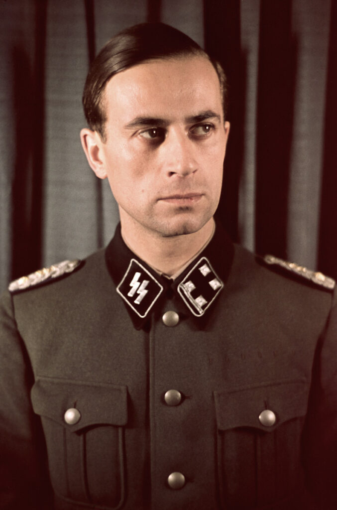 Es ist ein Farbporträt des jungen Karl Brandt. Er ist von vorne aufgenommen und guckt mit ernstem Blick nach rechts. Er hat die kurzen Haare streng zurück gekämmt. Er trägt die hochgeschlossene Uniform der SS.